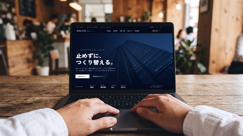 木目調のカフェで、ノートパソコンにコーポレートサイトを表示して操作している様子